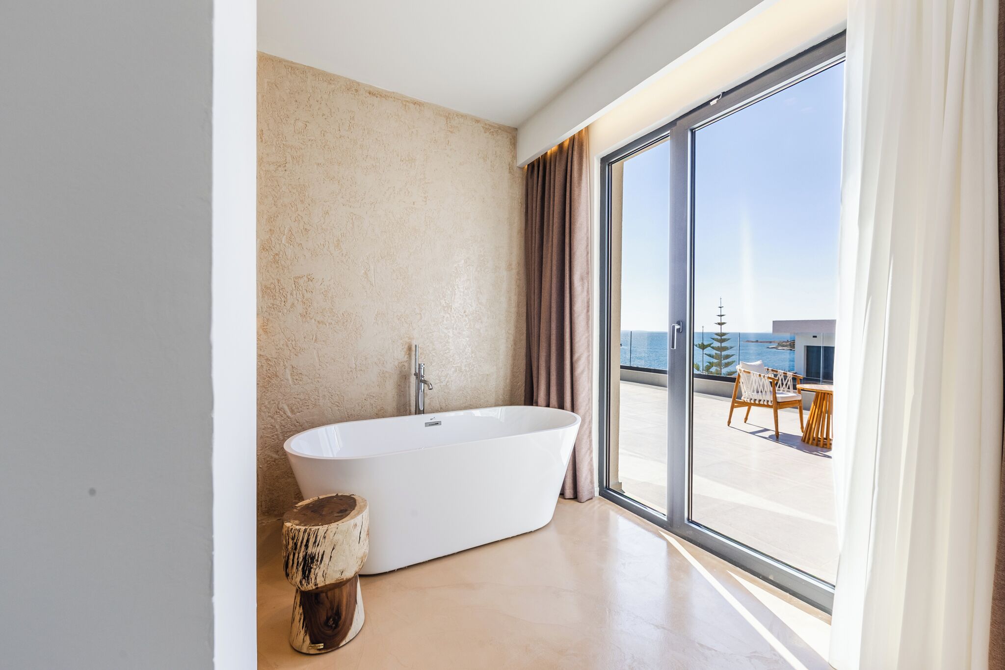 Un baño moderno con una bañera independiente, amplias puertas correderas de vidrio y vistas a un balcón con mobiliario exterior.
