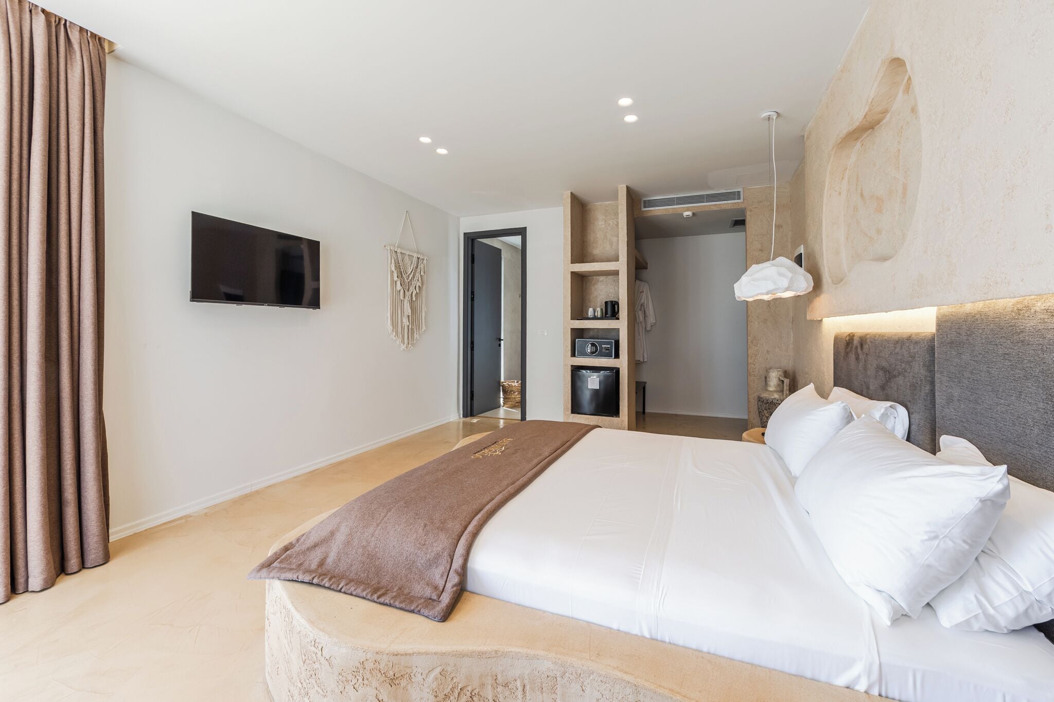 Een moderne hotelkamer met een groot bed, een flatscreen televisie, een houten hoofdbord en een inbouwkastje met een koffieapparaat.