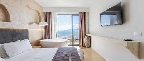 A modern hotel room with a large bed, a flat-screen TV, a bathtub, and a balcony with a view of the sea.