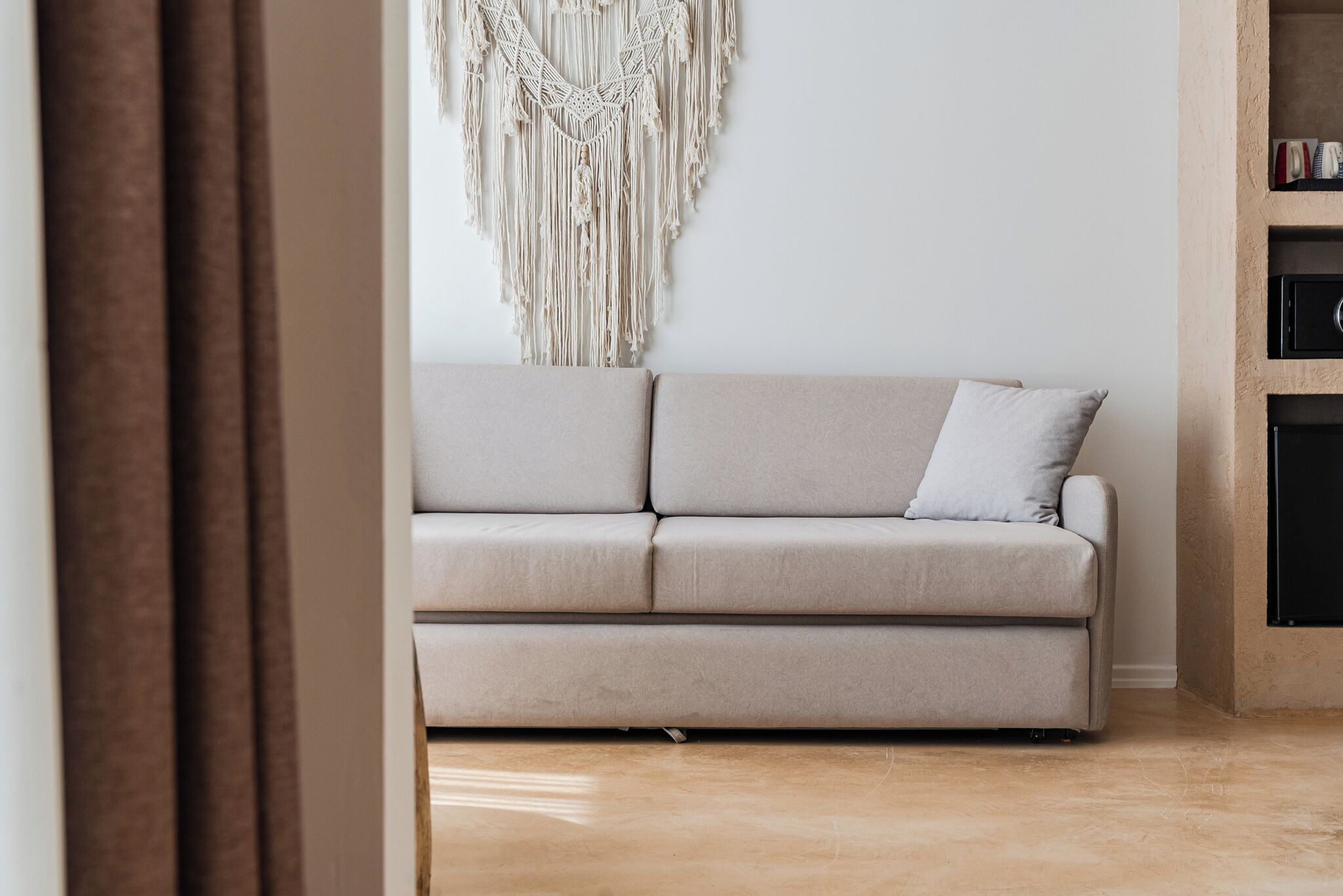 A neutral-toned sofa with a cushion, positioned against a wall with a macramé wall hanging.