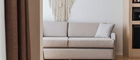 A neutral-toned sofa with a cushion, positioned against a wall with a macramé wall hanging.