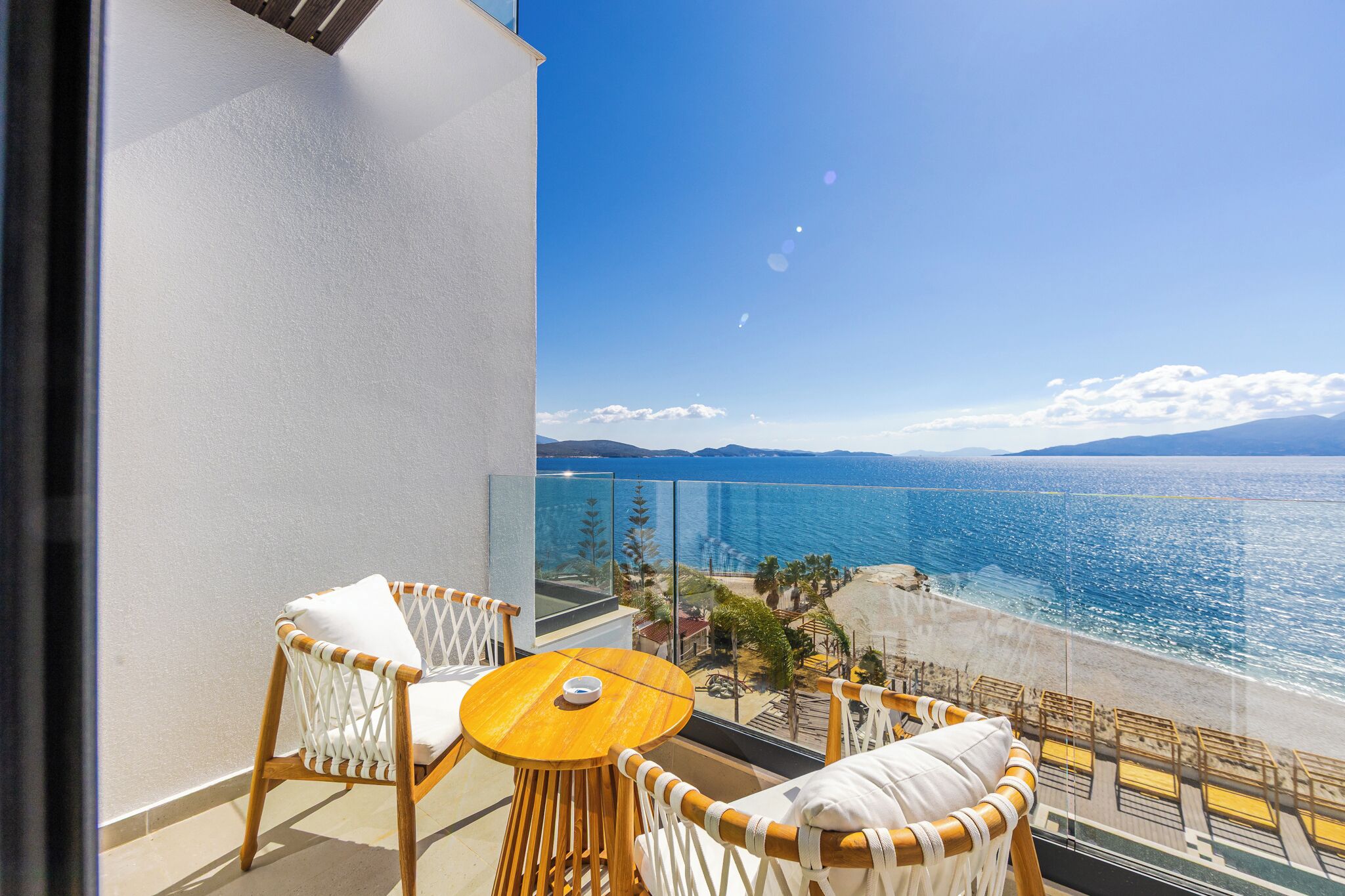 A balcony with wicker furniture, a round wooden table, and a view of the sea and beach.