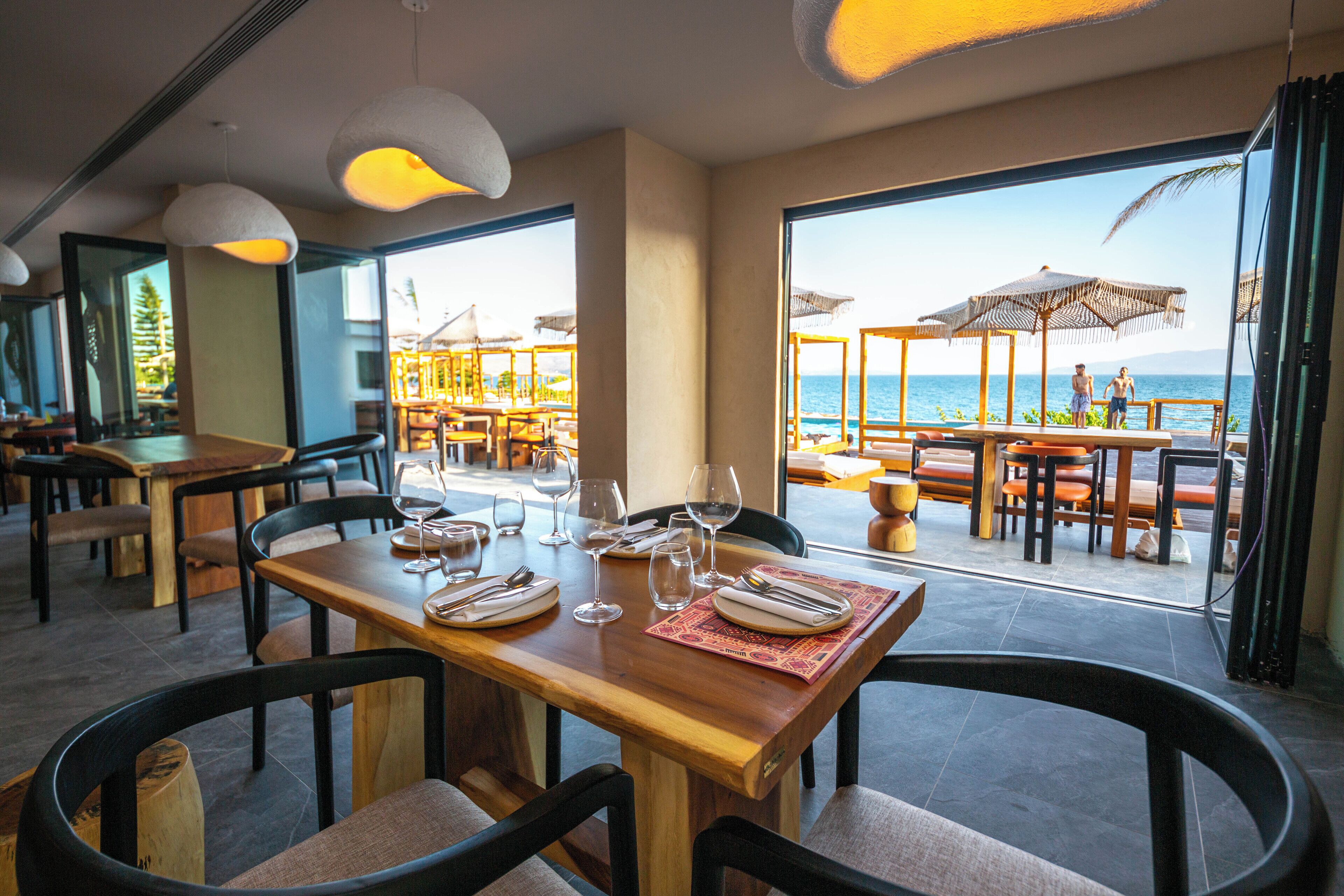 A restaurant with wooden tables and chairs, a view of the ocean, and umbrellas outside.
