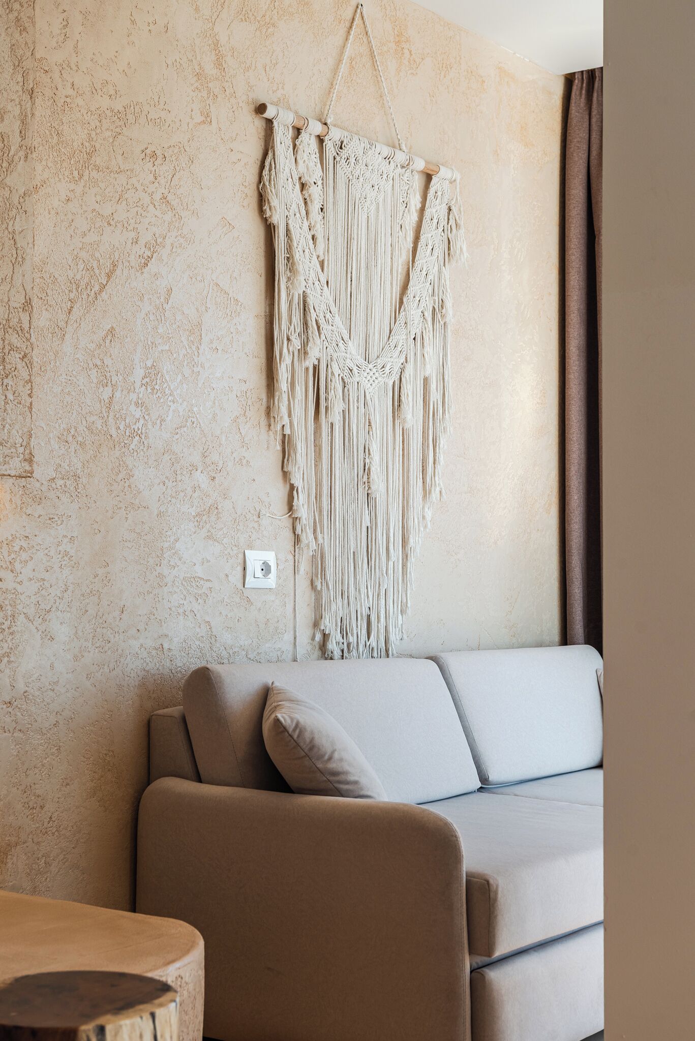 A room with a textured beige wall, a macrame wall hanging, a beige sofa, and a small wooden table.