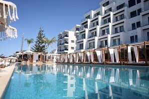 Une piscine entourée de chaises longues et de parasols, le tout bordé d’un bâtiment blanc doté de balcons.