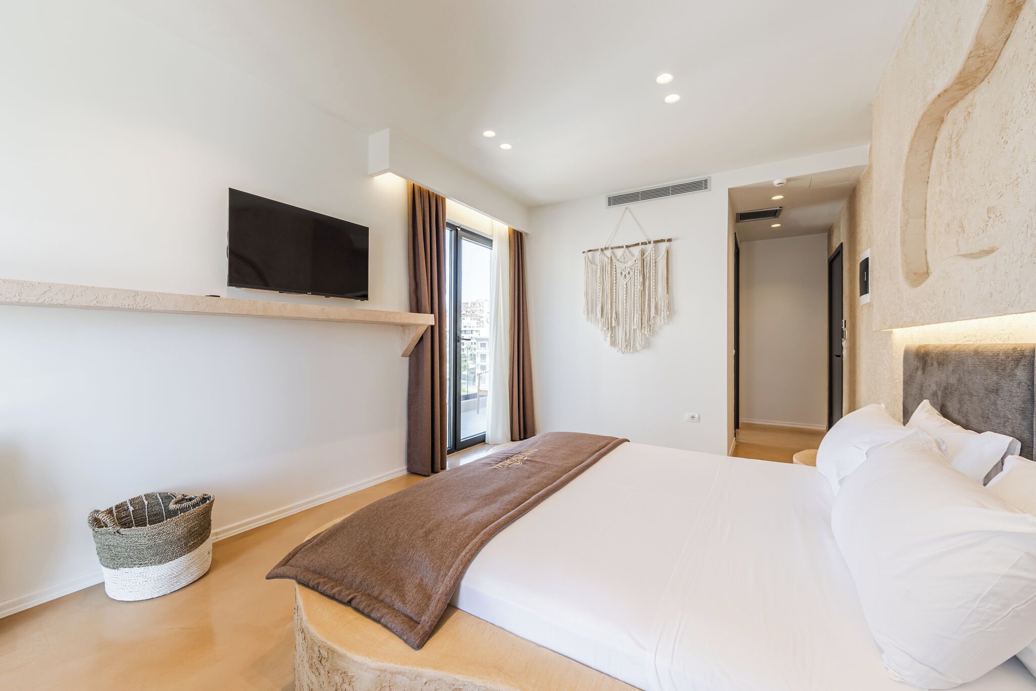 Habitación de hotel moderna con una cama grande, televisor montado, un pequeño rincón de descanso y vista a la ciudad desde la ventana.