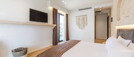 Habitación de hotel moderna con una cama grande, televisor montado, un pequeño rincón de descanso y vista a la ciudad desde la ventana.
