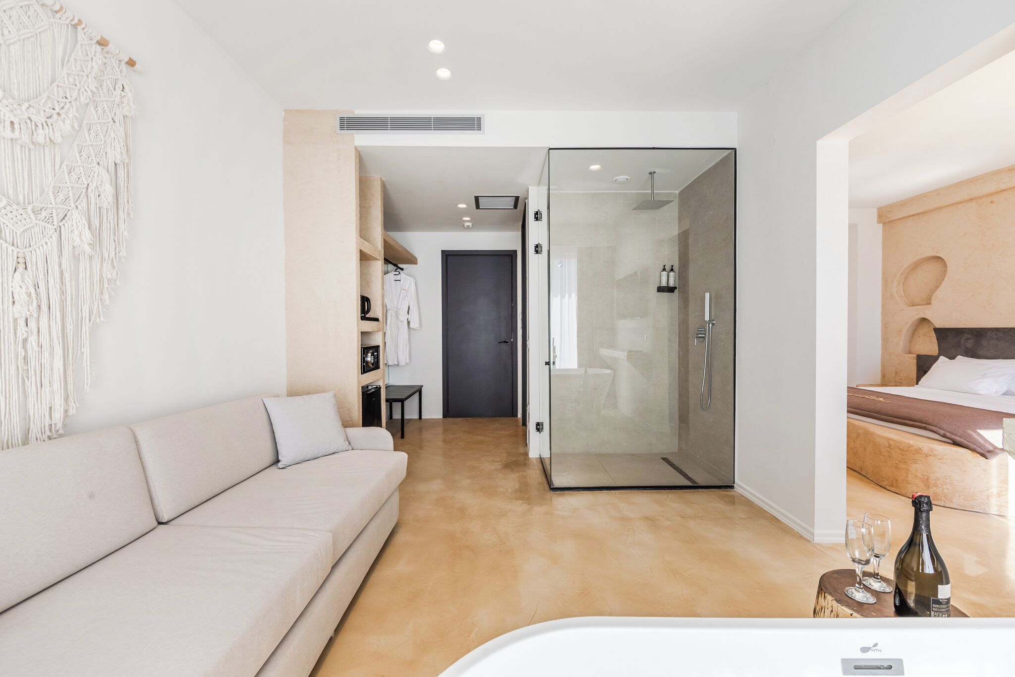 Een moderne hotelkamer met een beige bank, een glazen douchecabine, een houten hoofdbord en een fles champagne op een houten kruk.