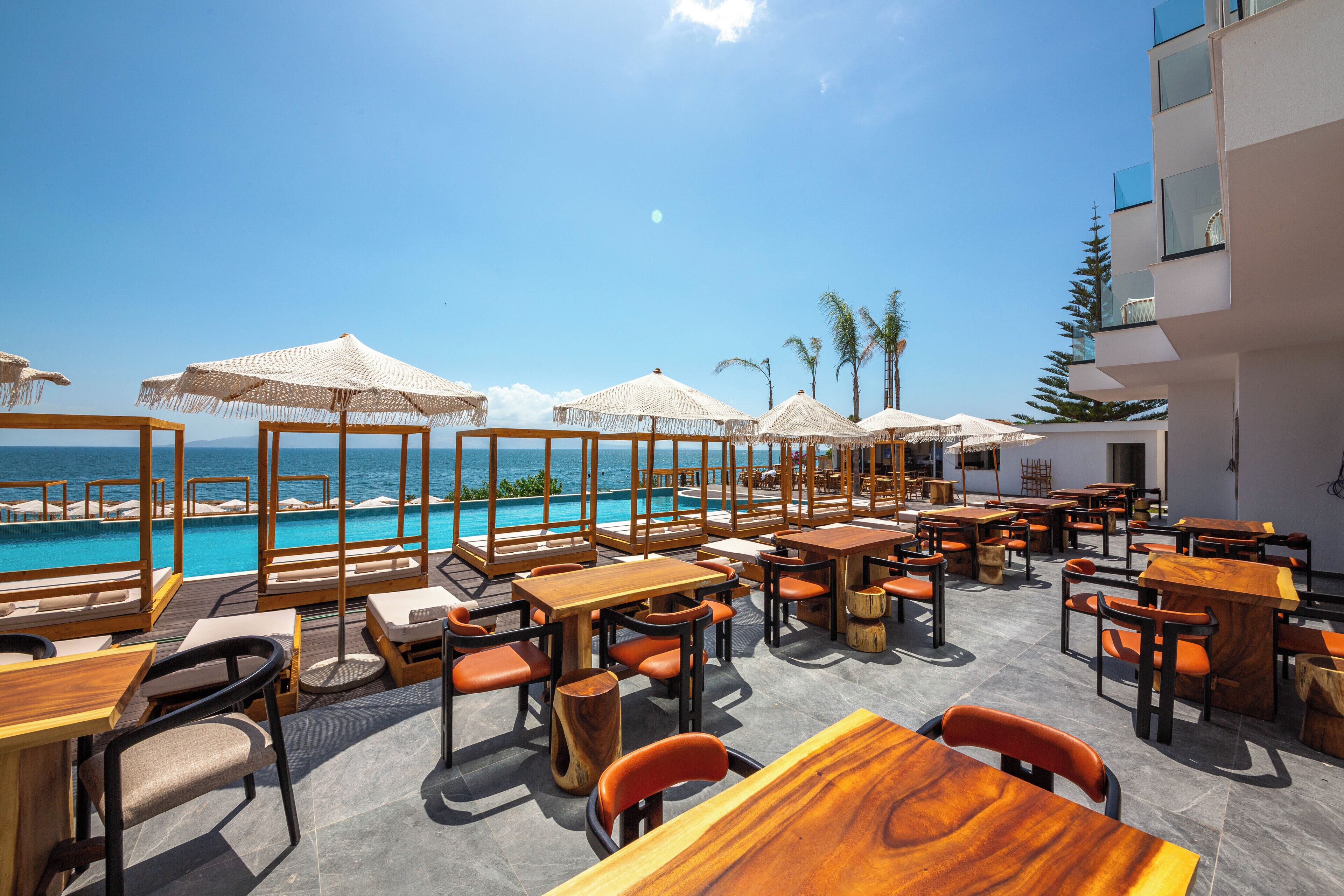 An outdoor seating area with wooden tables and chairs, umbrellas, and a pool view.