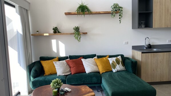 Een moderne woonkamer met een groene modulaire bank, meerdere kussens, een houten salontafel en een keuken met een spoelbak en kastjes.