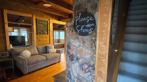 A cozy living room with a stone accent wall, a sign requesting guests to remove their shoes, a wooden staircase, and a comfortable sofa. - Family Friendly Log Cabin Nestled in Blue Ridge Foothills (Stuarts Draft)