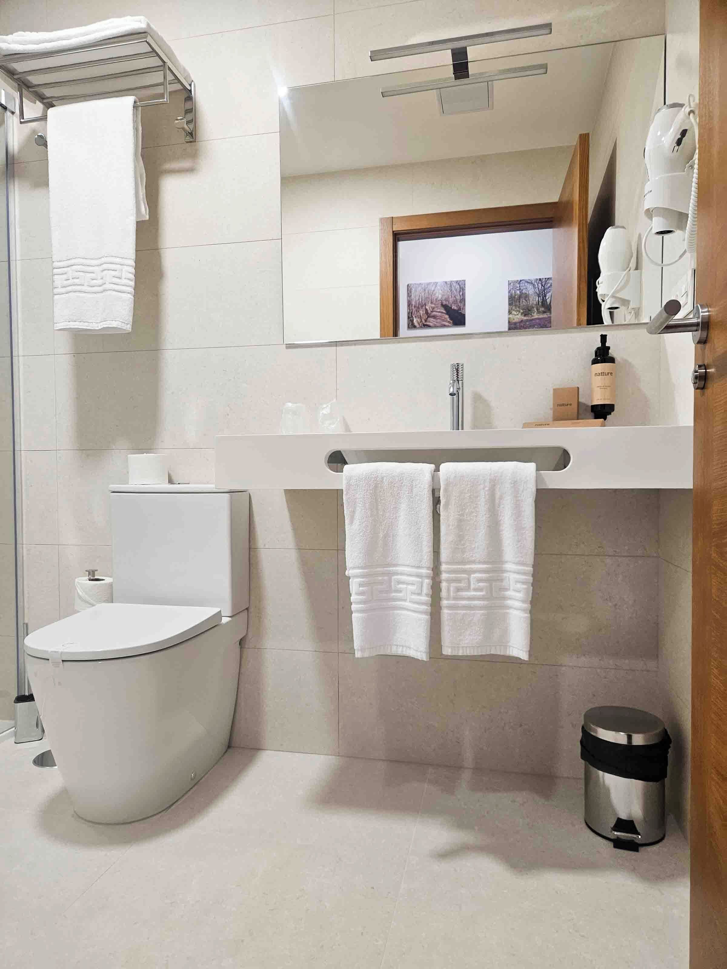 Een moderne badkamer met toilet, hygiëneslot en wastafel. Twee handdoeken hangen aan de muur, er is een prullenbak en een spiegel met een sierlijst.