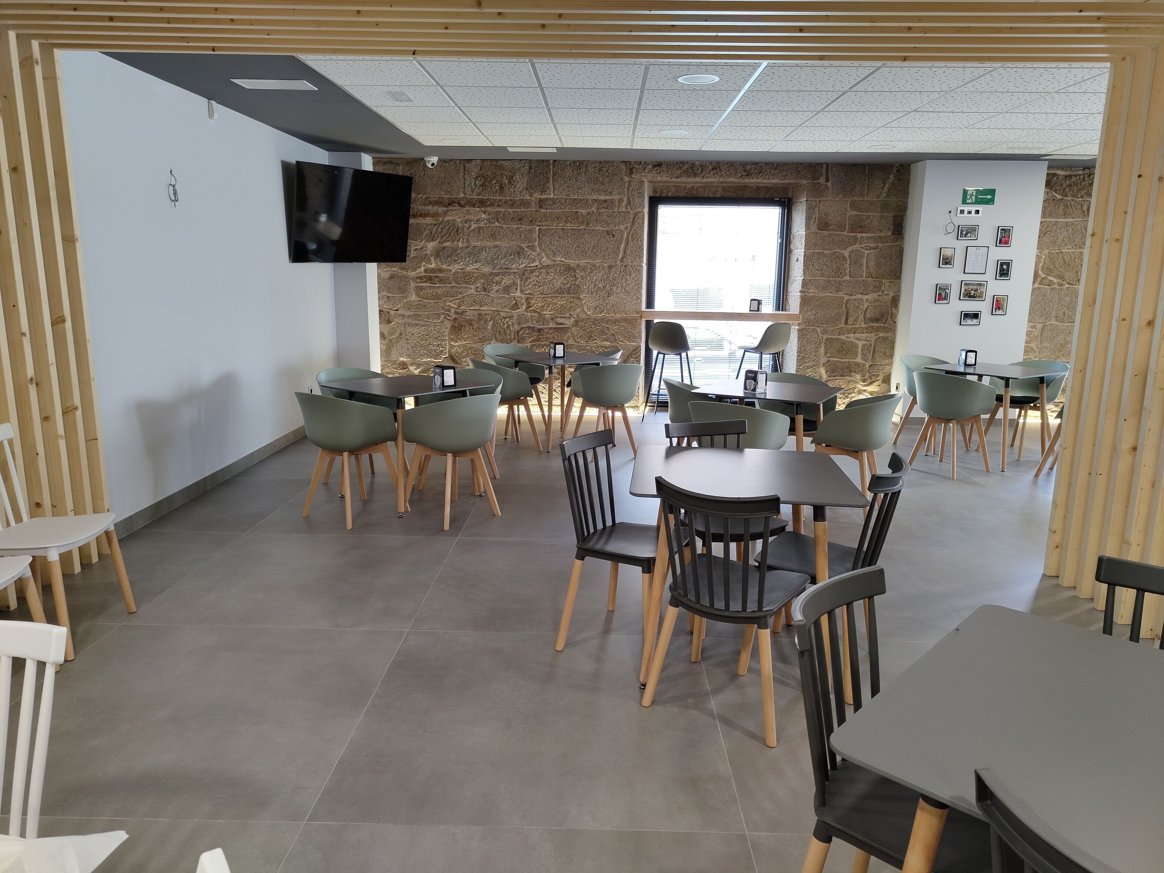 A modern dining area with multiple tables and chairs, a stone wall, a television mounted on the wall, and a window providing natural light.