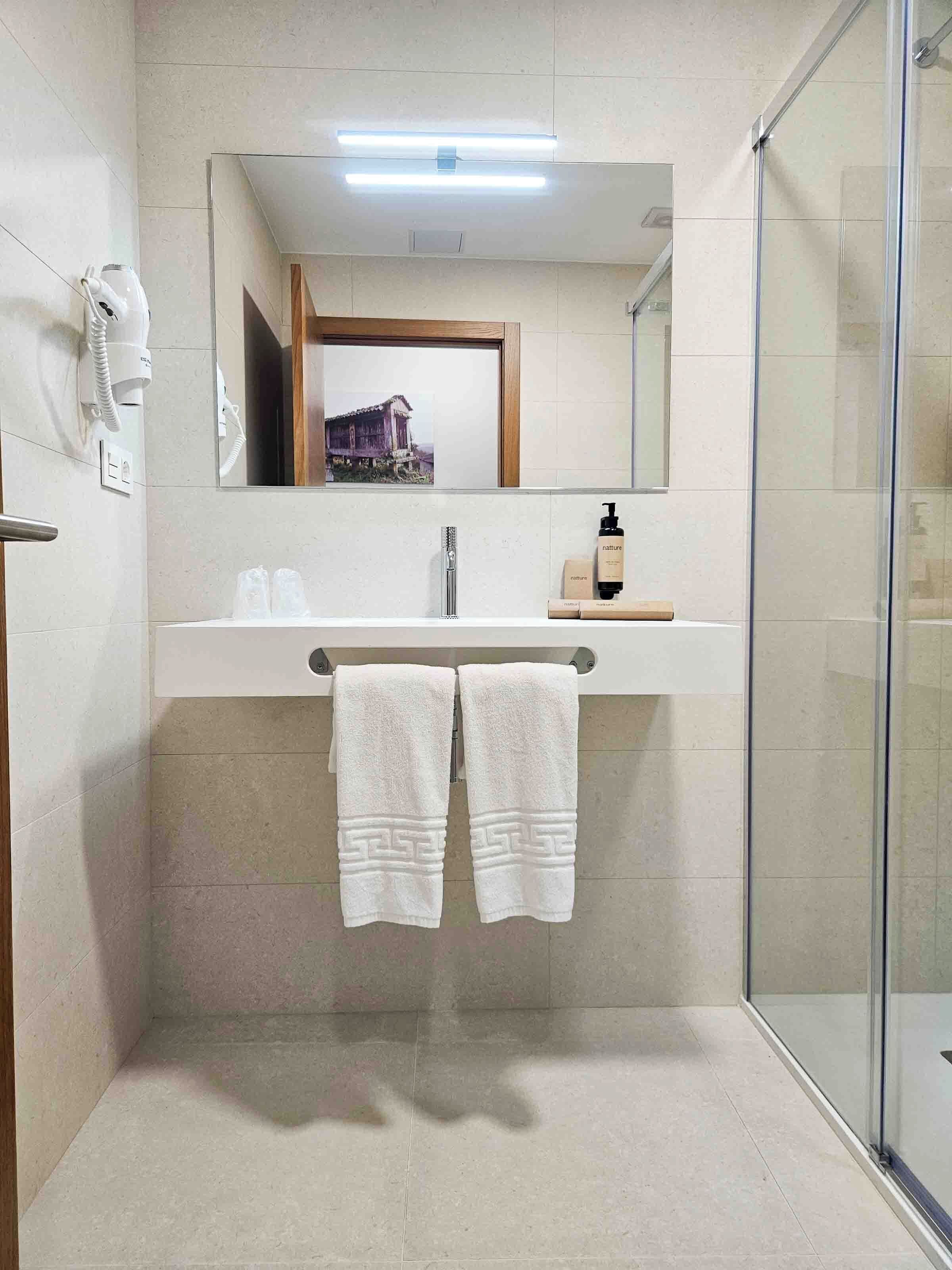 Een moderne badkamer met een douchecabine van glas, een wit wastafeltje met een spiegel erboven en twee handdoeken aan een rek.