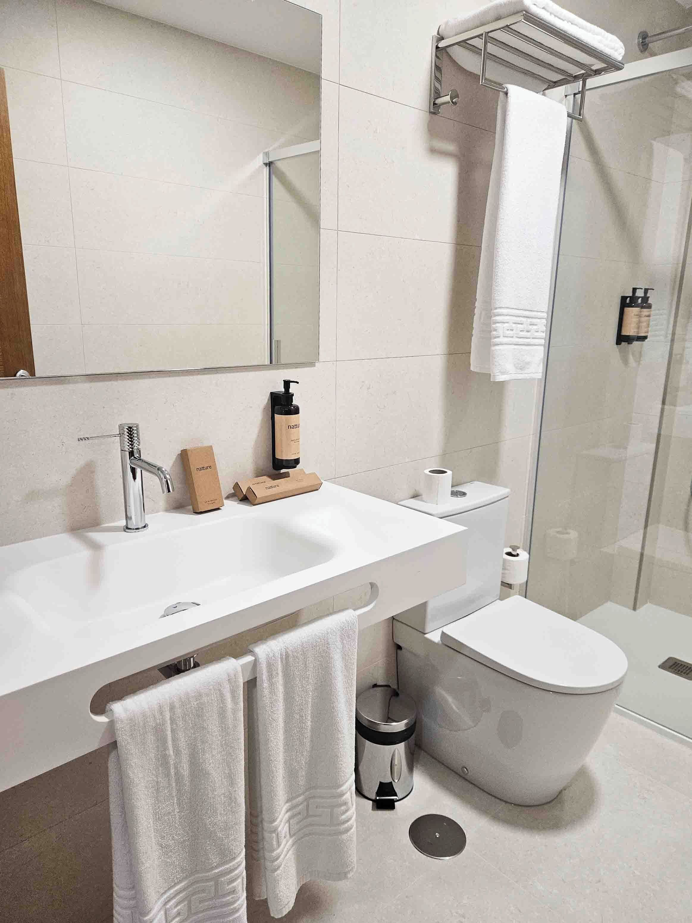 Et moderne badeværelse med en hvid vask, spejl og toilet. Der er et bruseområde med en glasdør og en bøjle til håndklæder over vasken.