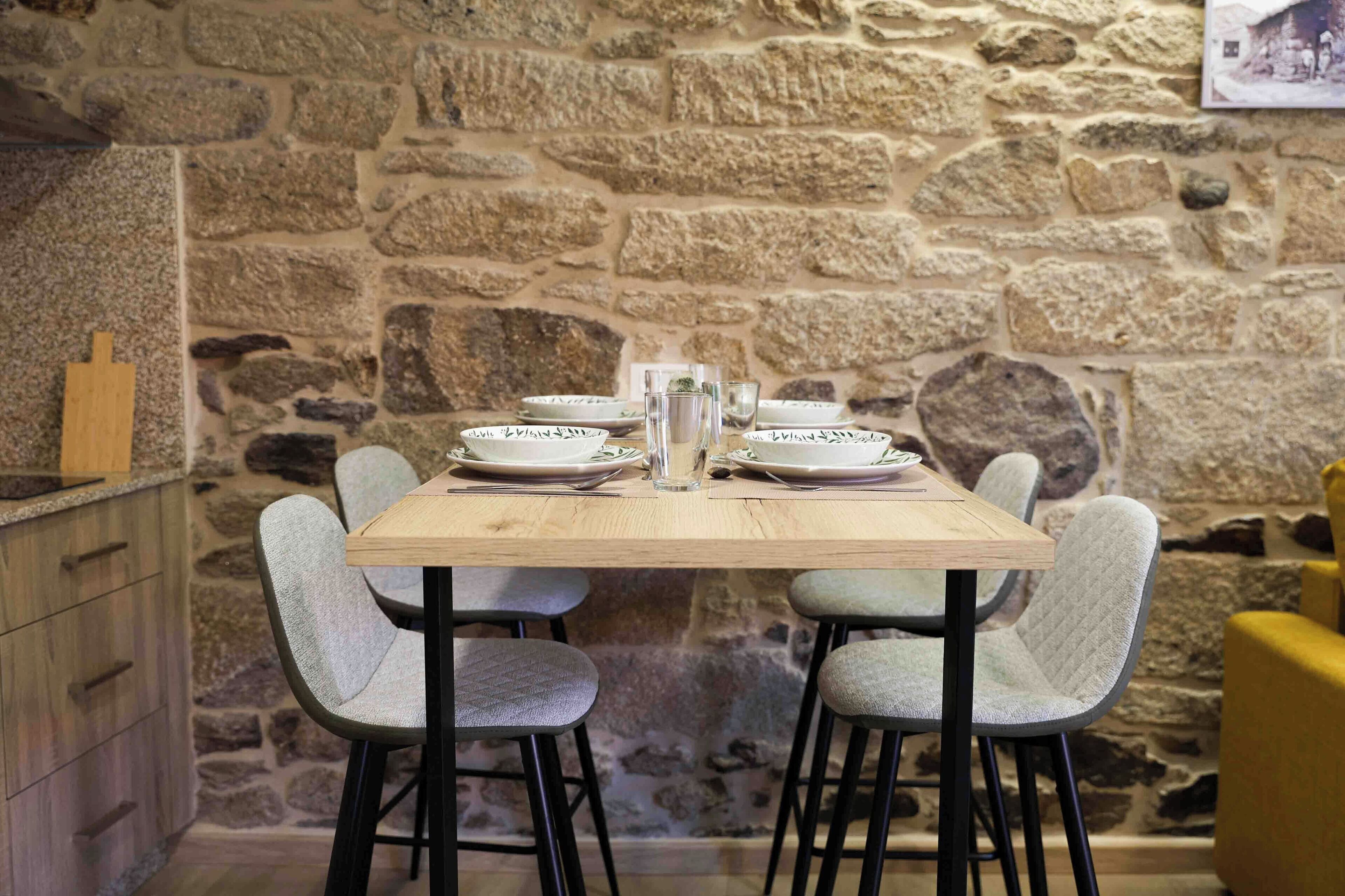Mesa de jantar de madeira com quatro cadeiras cinzas estofadas, posta com pratos, copos e um jarro. Uma parede de pedra e uma cadeira amarela estão ao fundo.