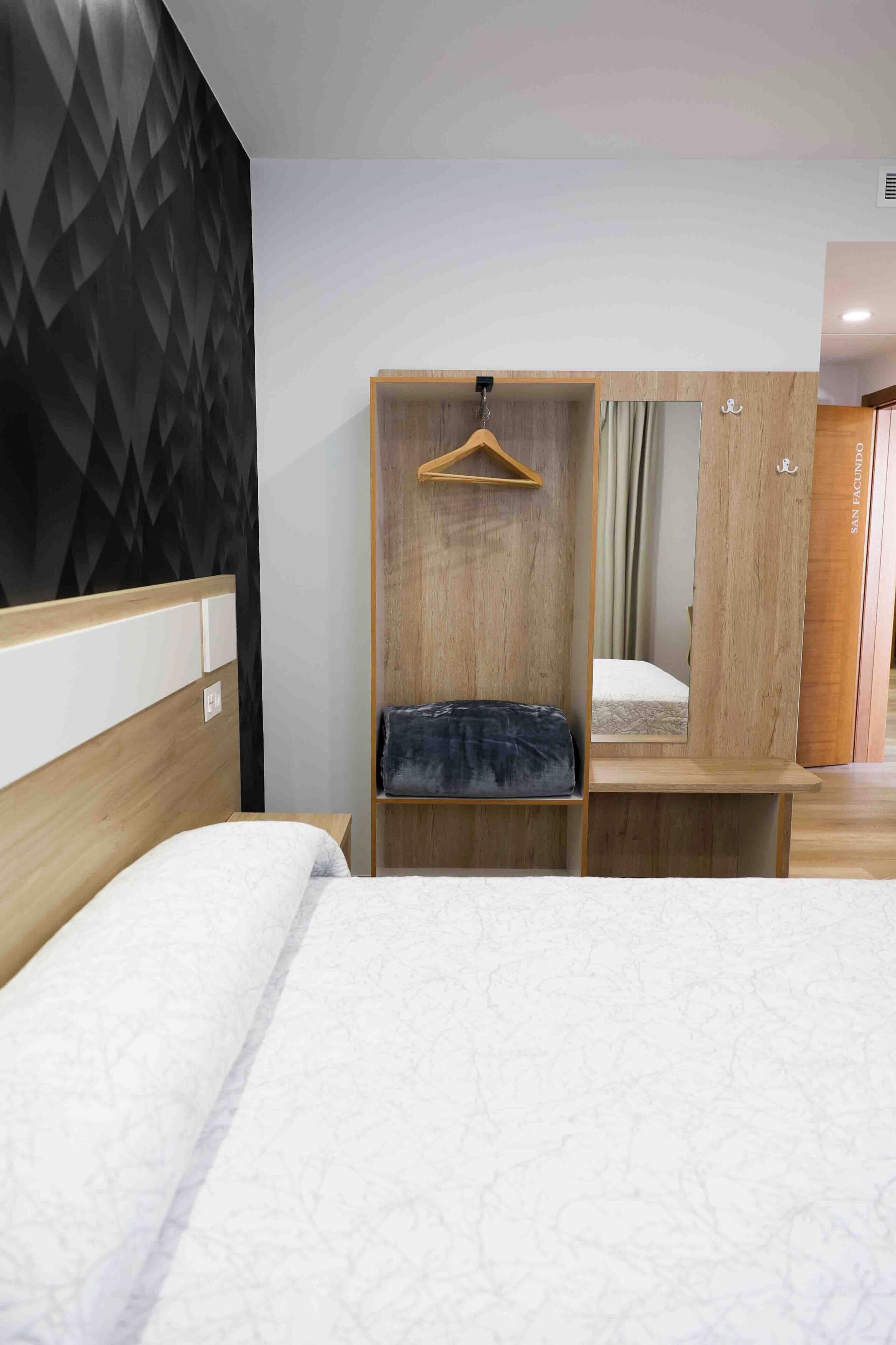 A hotel room with a bed, a wooden wardrobe, a hanging coat rack, and a mirror.