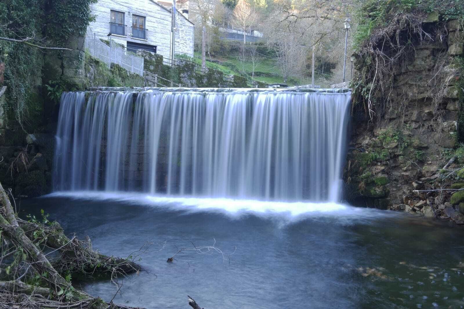 Ein Wasserfall stürzt über einen Felsvorsprung in einen Pool, umgeben von viel Grün und einem Steinhaus.