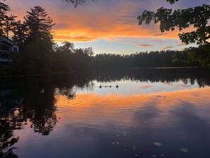 A tranquil lakeside scene with a house on the left and a sunset reflecting on the water.