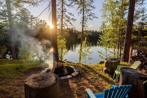 A campfire surrounded by trees, a lake, and a wooden deck with chairs.