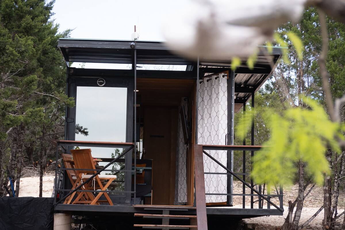 Una cabaña moderna con una terraza de madera, una silla y una escalera que lleva a la entrada.