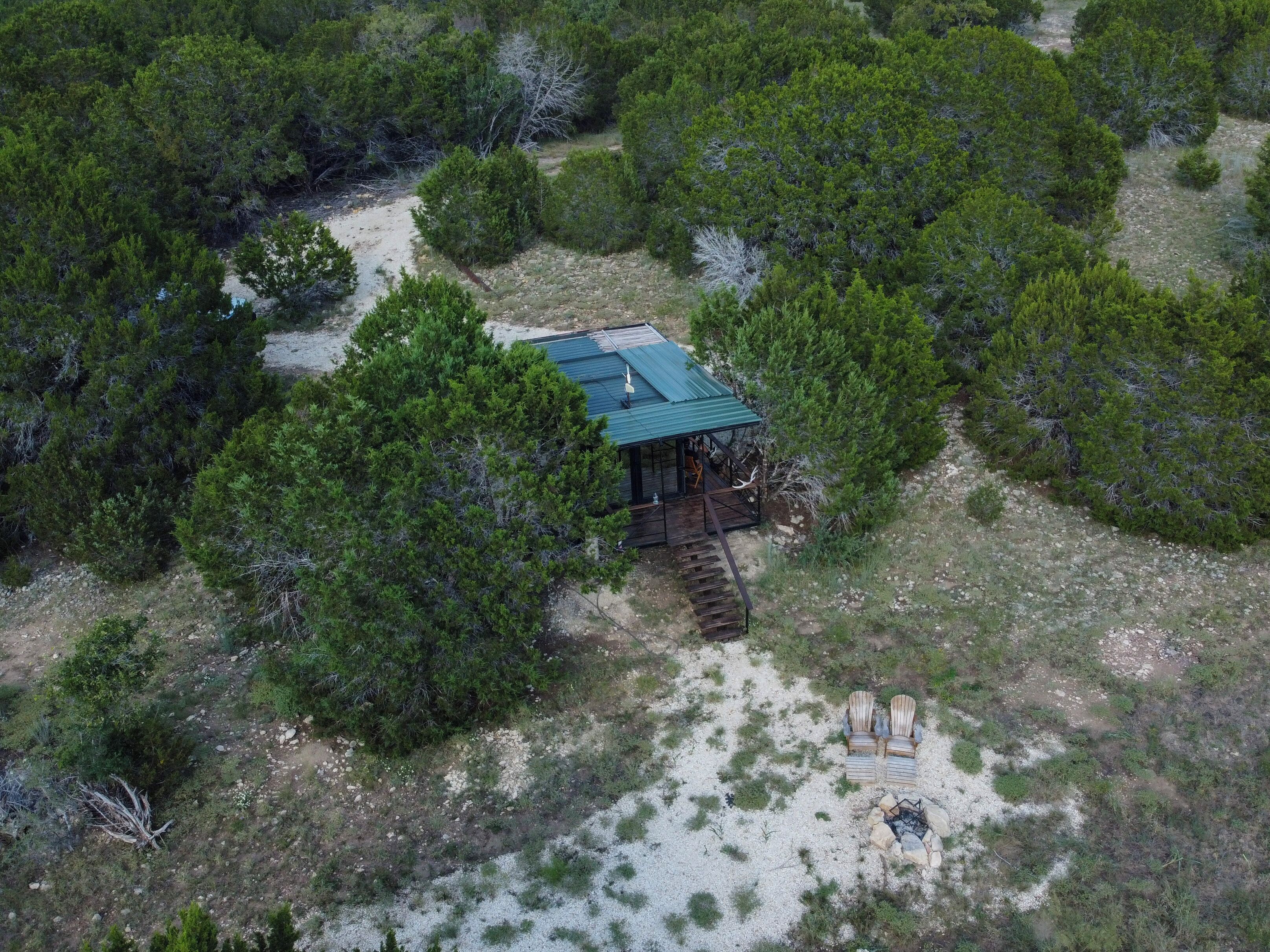 Une cabane isolée, entourée d’une forêt dense, avec un chemin de gravier menant à elle et un petit foyer à proximité.