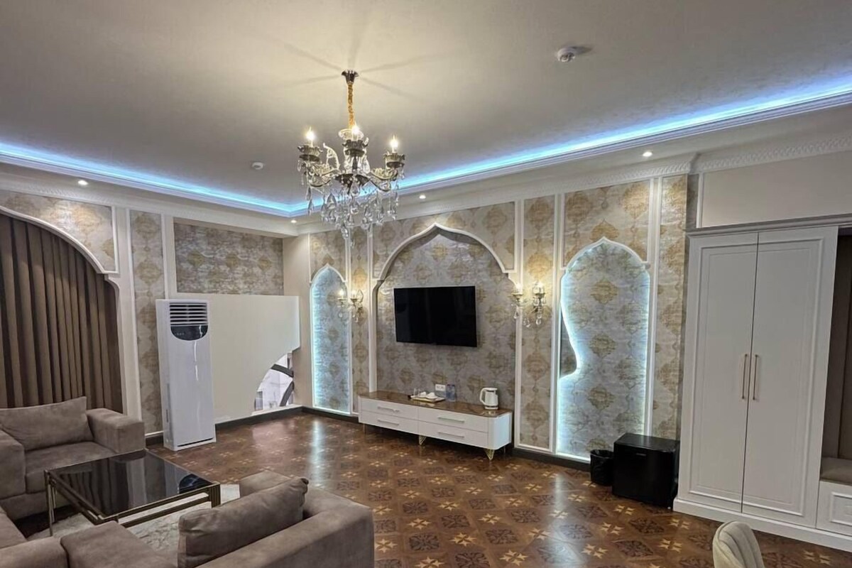 A spacious living room with a modern chandelier, a flat-screen TV mounted on a decorative wall, and a patterned floor.