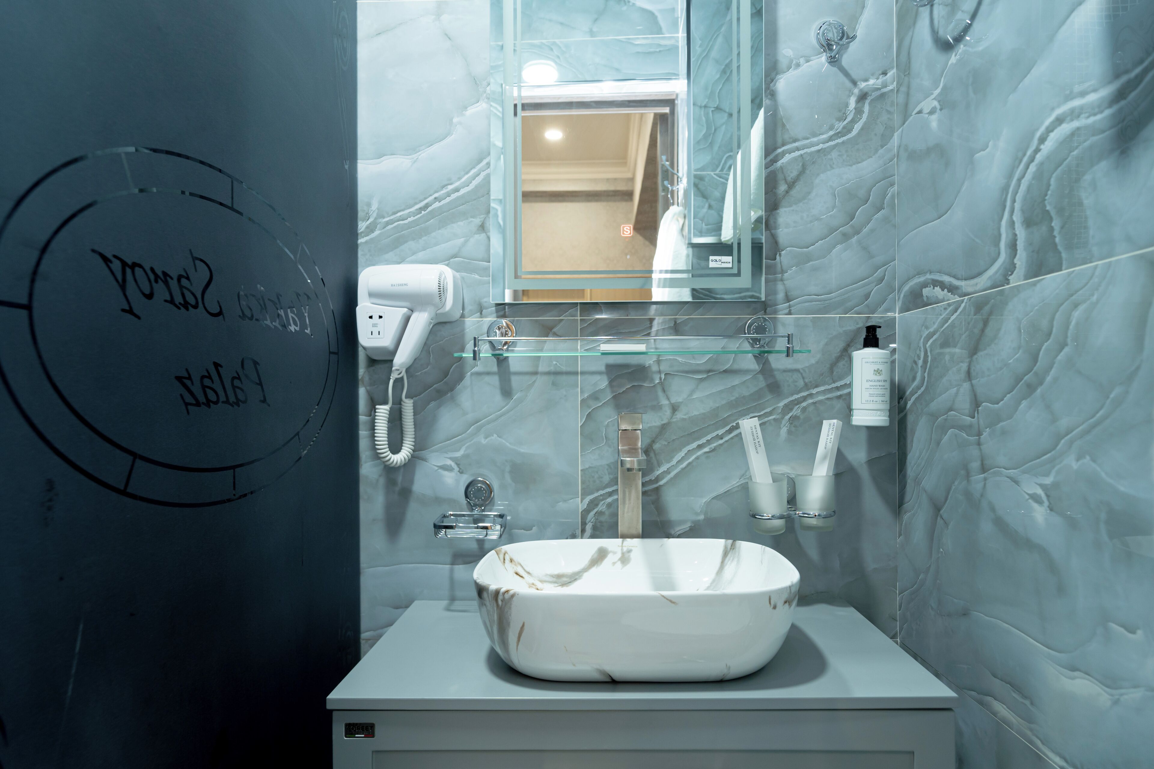 A bathroom with a marble wall, a large sink, a mirror, a hairdryer, and toiletries.