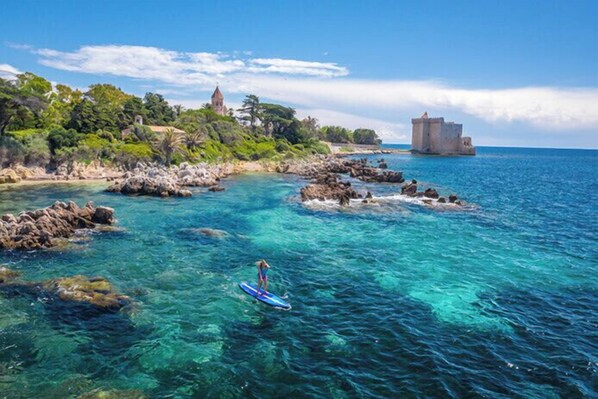 A coastal scene with clear turquoise waters, rocky outcrops, and a person paddleboarding.