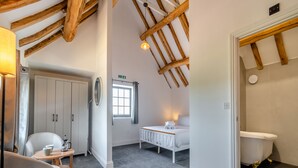 Een kamer met een hoge plafond, zichtbare houten balken, een wit toilet, een bed met een witte kap, een klein rond tafeltje met een stoel, en een raam met uitzicht op de omgeving.
