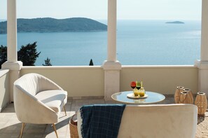 Un balcon avec vue sur la mer, une table ronde avec des boissons, un fauteuil blanc et des vases en osier.