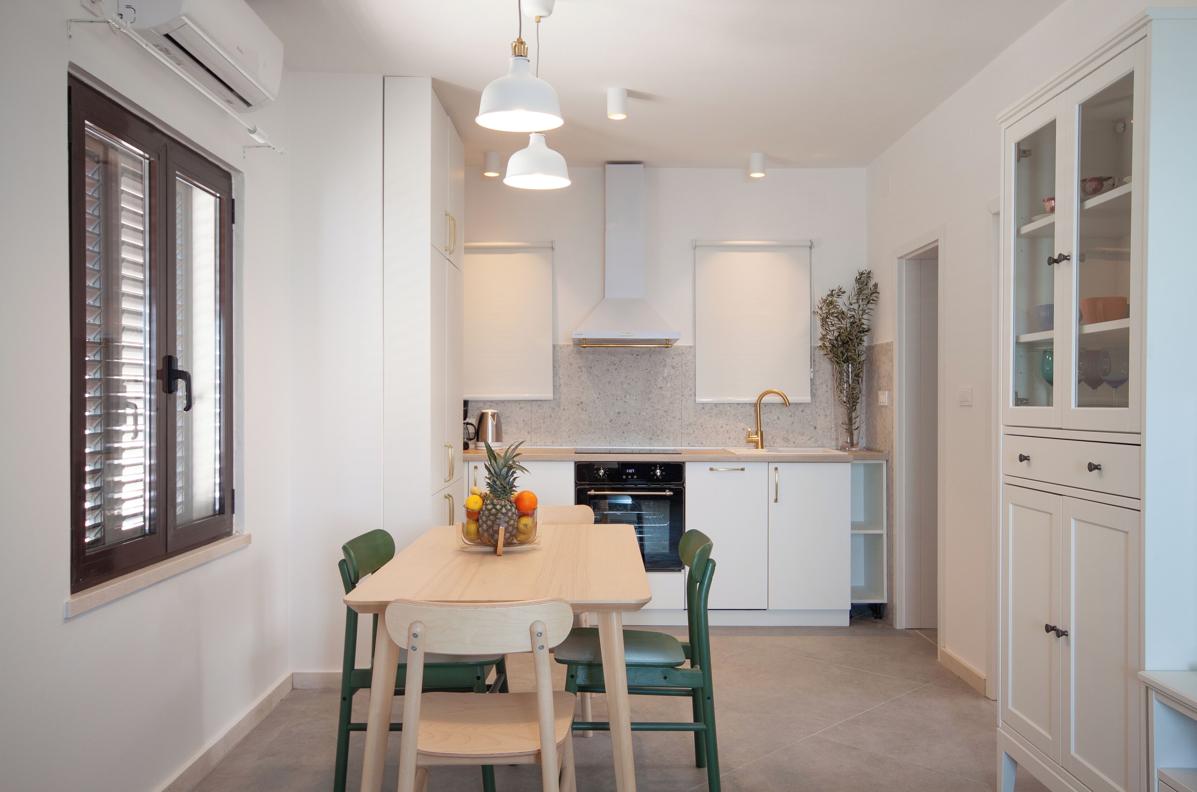 Una cocina moderna con armarios blancos, una mesa de comedor de madera y dos sillas. Hay un frutero en la mesa y una ventana con persianas a la izquierda.