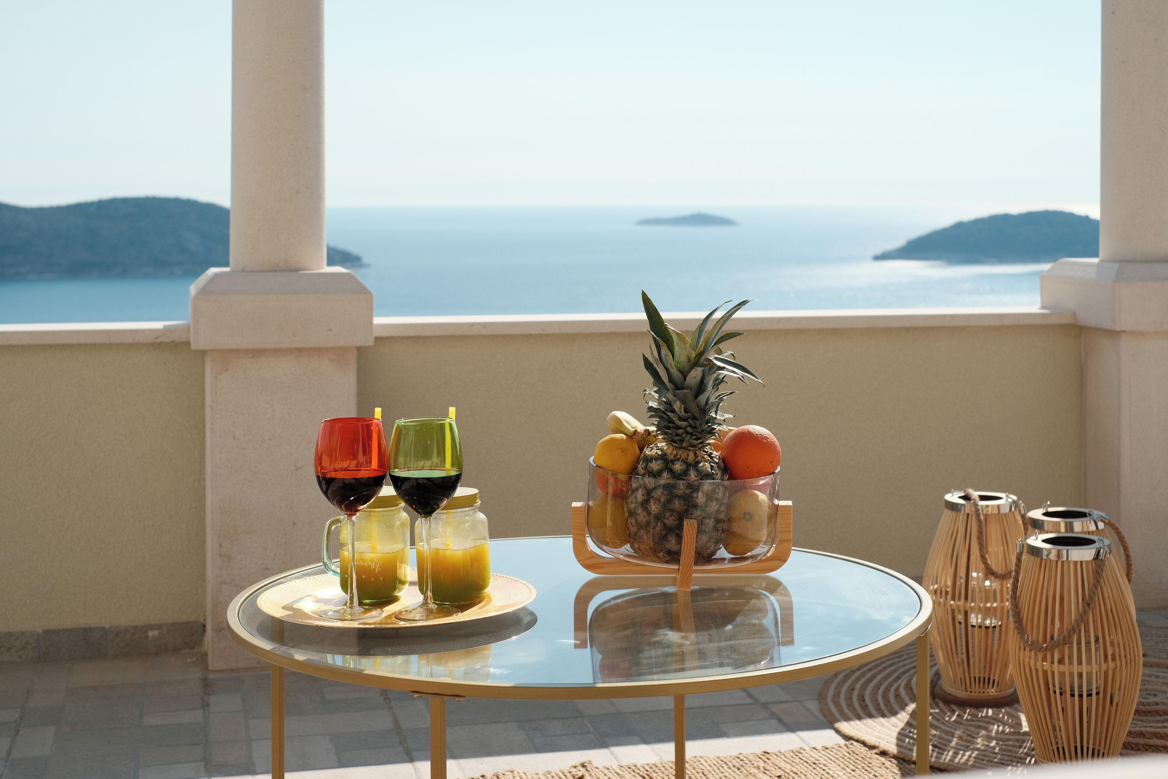 A round glass table with a fruit bowl, two glasses of juice, and two candles on a balcony overlooking the sea.