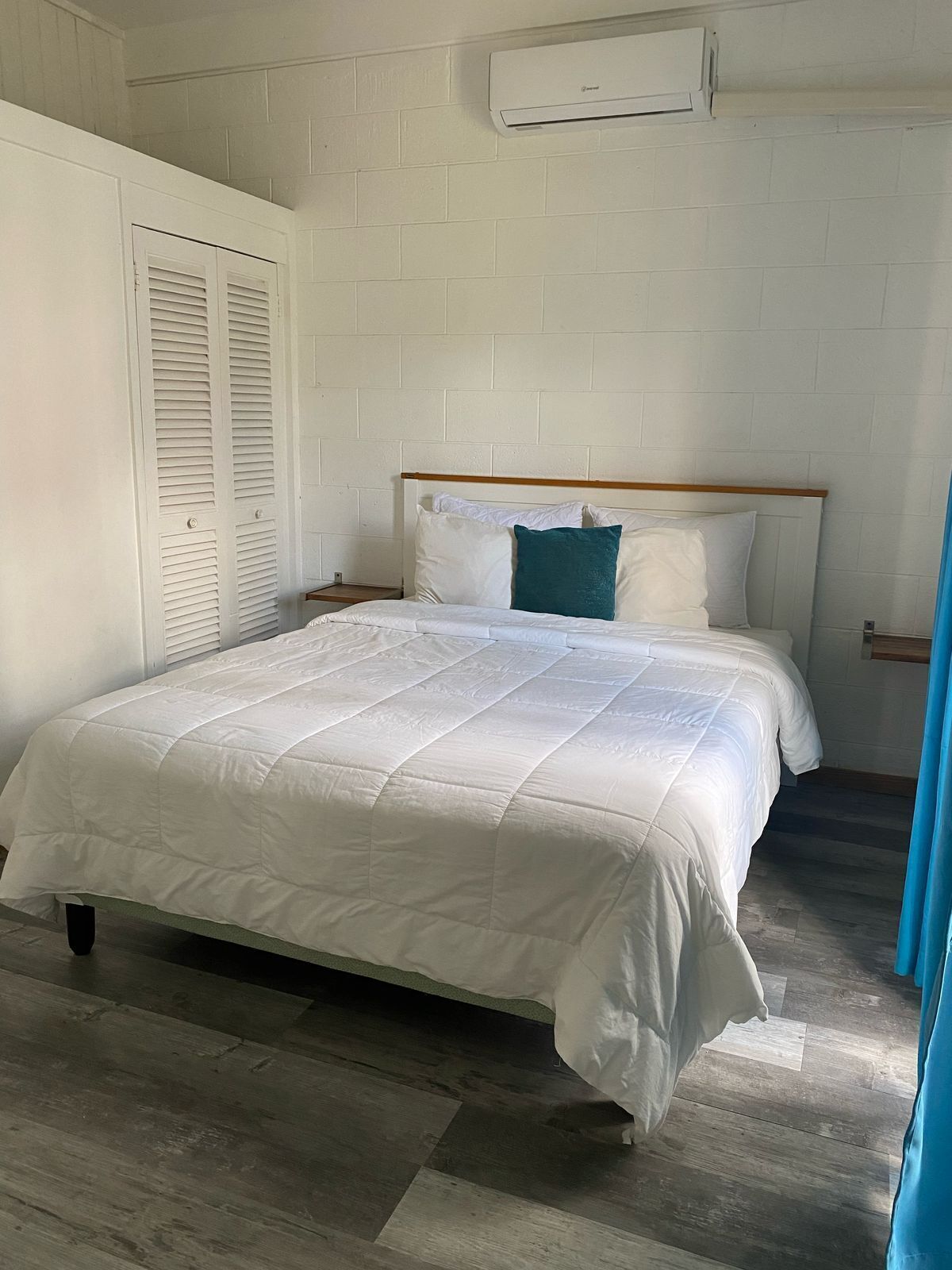 Ein Schlafzimmer mit einem weißen Bett, einem hölzernen Kopfteil, einer weißen Wand und einem Fenster mit blauen Vorhängen.