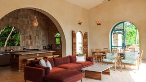 A spacious living area with a red sofa, wooden dining table, and chairs, a kitchen with a bar, and large windows offering views of greenery. - Surf, Sun, Serenity! Luxury Private NEW Villa w/ Pool, Ocean and Valley Views.  (San Juan del Sur)