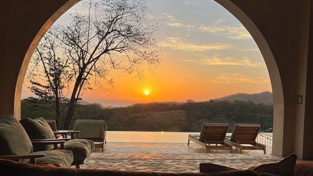 A room with a large arched window overlooking a sunset over a body of water, with lounge chairs and a small table. - Surf, Sun, Serenity! Luxury Private NEW Villa w/ Pool, Ocean and Valley Views.  (San Juan del Sur)