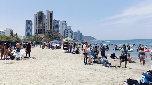 Una spiaggia affollata con persone che prendono il sole e nuotano, e una città sullo sfondo.