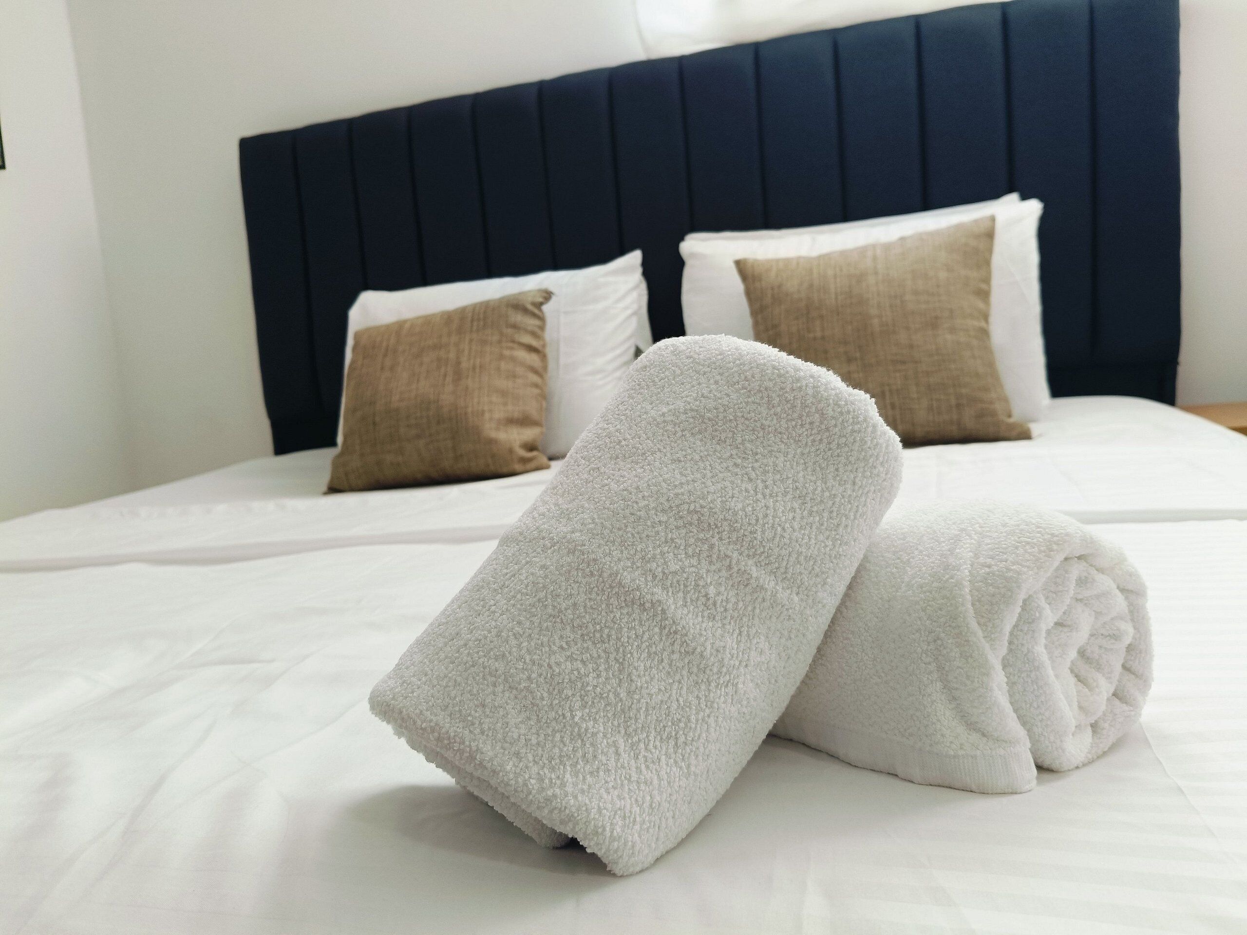Een net opgemaakt bed met witte lakens, twee opgerolde handdoeken, een donker hoofdbord en twee kussens met verschillende texturen.