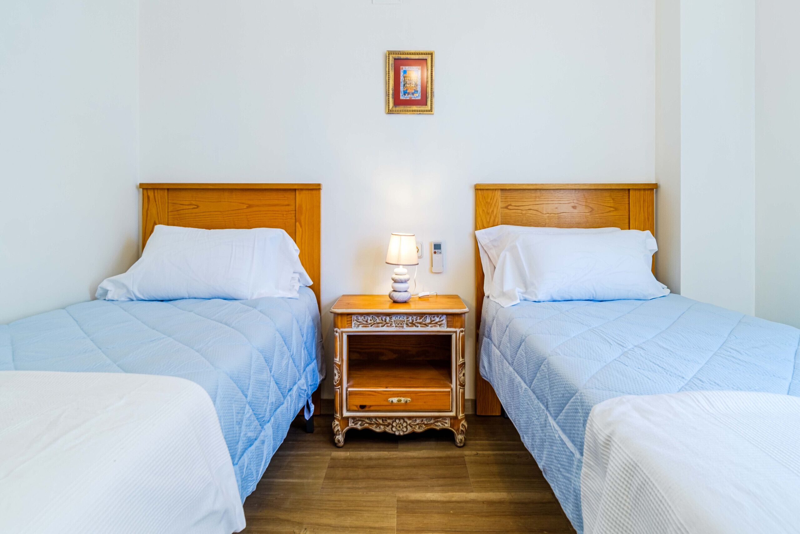 Una habitación con dos camas, cada una con cabecero de madera y almohada blanca. Entre las camas hay una mesita de noche con una lámpara y un cajón pequeño. Encima de la mesita de noche, un cuadro enmarcado cuelga de la pared.