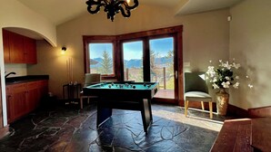 Une table de billard dans une pièce dotée d’un sol en pierre, de meubles en bois, d’un lustre et offrant une vue sur l’extérieur grâce à une grande fenêtre.