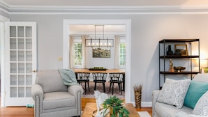 A living room with a sofa, a coffee table, and a bookshelf. There is a dining area with a table and chairs in the background. - Discover university charm. Your 4-bedroom retreat, an 8-min drive to Notre Dame. (South Bend)