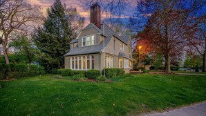 A two-story house with a prominent chimney, surrounded by a well-maintained lawn and trees. - Discover university charm. Your 4-bedroom retreat, an 8-min drive to Notre Dame. (South Bend)