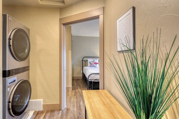 Un área de lavandería con lavadora y secadora, una mesa de madera, una cama con almohadas y un letrero en la pared.