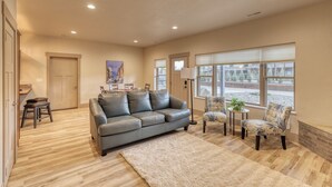 A spacious living room with a grey leather sofa, a small table with chairs, a large window with blinds, and a door leading to another room. - Mt Rushmore Retreat hosted by Rockerville Gold Town Lodging • 8 Min Drive | 2 Bedroom Sleeps 6- Fireplace (Rockerville)