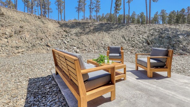 Eine Holzbank und zwei Stühle auf einer Beton-Terrasse, im Hintergrund ein felsiger Hang und Kiefern.