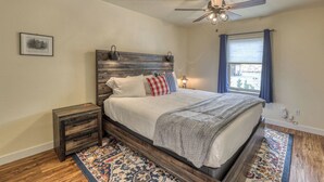 A bedroom with a wooden headboard, a bed with white bedding, a nightstand, a floor lamp, a ceiling fan, a window with curtains, and a patterned rug. - Homestake Haven Hosted by Rockerville Gold Town Lodging-Black Hills Base Camp-2 Bedroom, King Suite (Rockerville)