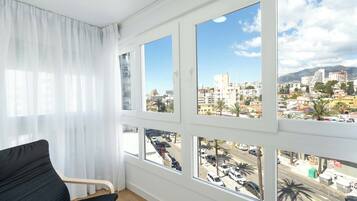 Una habitación con una silla negra, ventanas amplias y vistas a un paisaje urbano con edificios y palmeras.