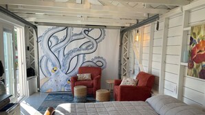A bedroom with a bed, a red armchair, a small table, and a wall mural. - Similk Beach View Studio for two! 
Central HUB to San Juans and Skagit Valley! (Anacortes)