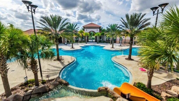 A swimming pool with a water slide, surrounded by palm trees and a building with a red-tiled roof. - Newly Remodeled Disney Vacation Home (Davenport)