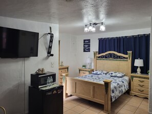 A bedroom with a bed, a television, a refrigerator, a bedside table with a lamp, and a ceiling light fixture.